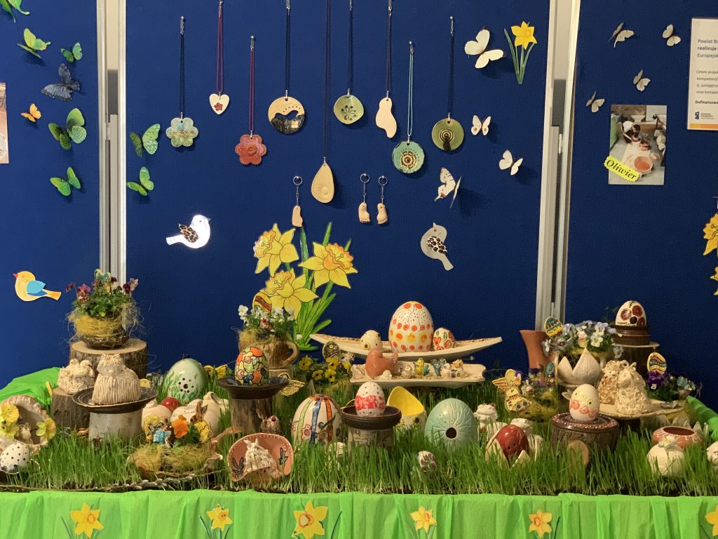 Ozdoby wielkanocne z ceramiki ustawione na stole w trawniku z młodego owsa. za stołem na niebieski tle wiszą ceramiczne ptaszki, jajka, motyle.