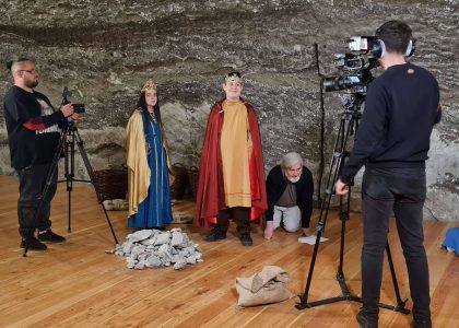 dziewczyna i chłopiec ubrani w stroje średniowieczne stoją na tle solnej ściany. Obok stoi mężczyzna z kamerą.