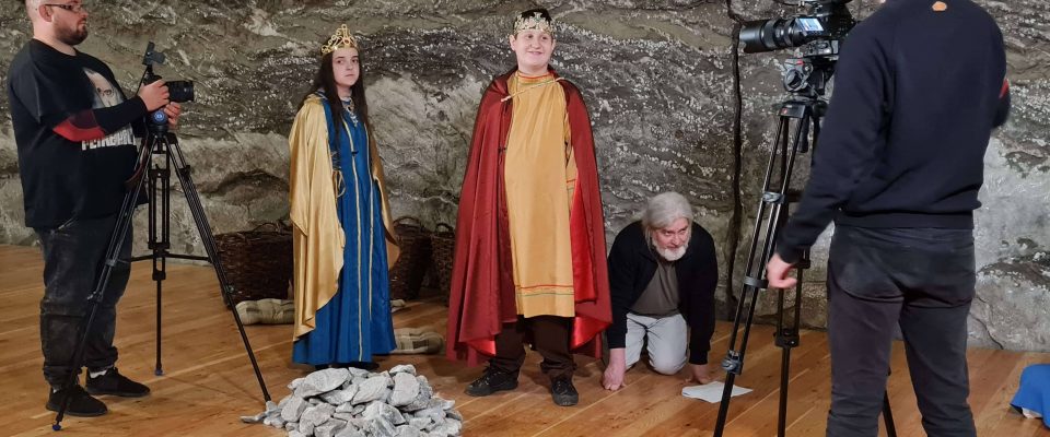 dziewczyna i chłopiec ubrani w stroje średniowieczne stoją na tle solnej ściany. Obok stoi mężczyzna z kamerą.