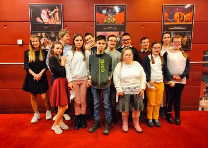 Grupa młodzieży pozuje do zdjęcia na tle czerwonej ściany. z plakatami sztuk do wystawienia.