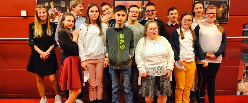 Grupa młodzieży pozuje do zdjęcia na tle czerwonej ściany. z plakatami sztuk do wystawienia.