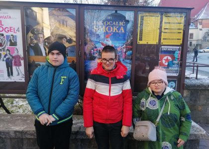 Troje dzieci ubranych w kurtki stoi obok plakatu zapowiadającego premierę kinową filmu.