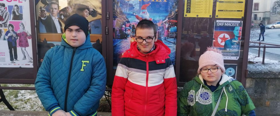 Troje dzieci ubranych w kurtki stoi obok plakatu zapowiadającego premierę kinową filmu.