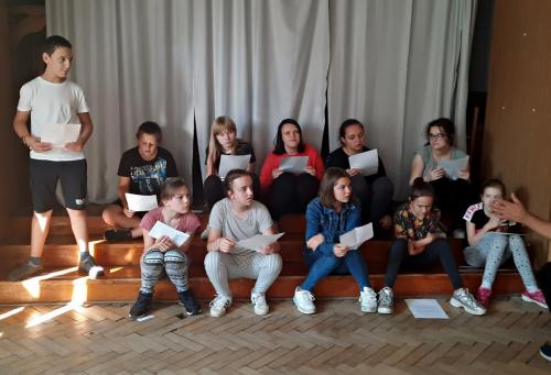 Grupa młodzieży sedzi na schodach sceny, w dłoniach trzymają kartki.