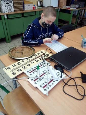 Chłopiec w maseczce siedzi przy stoliku, na którym leżą duże termometry i laptop.