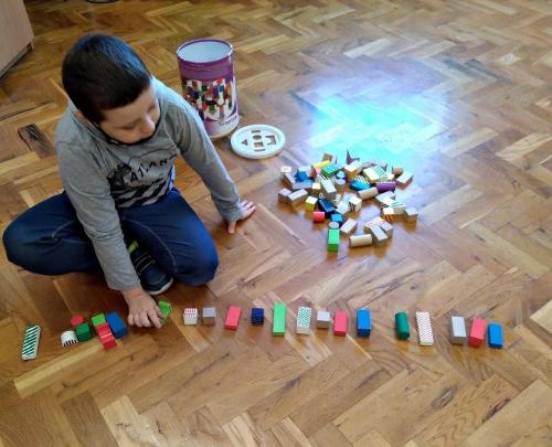 Chłopiec siedzi na podłodze i układa w ciągu  kolorowe, drewniane klocki.